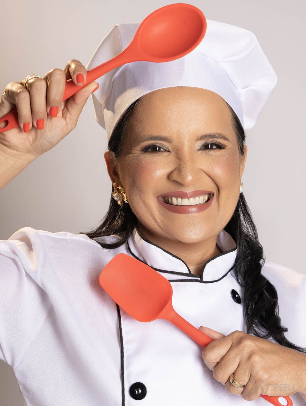 Ensaio de marca pessoal: chef de dólmã branca e chapéu segurando duas espátulas vermelhas, sorrindo, sobre fundo claro