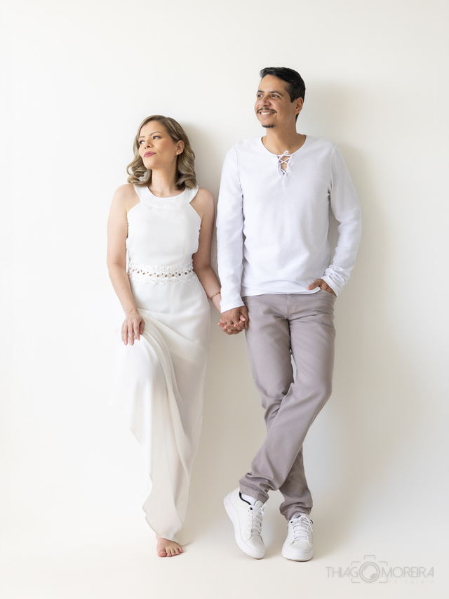 Casal à espera de um bebê, os dois vestidos de branco, caminhando de mãos dadas sobre fundo liso branco