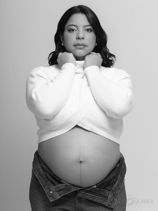 Retrato de gestante em preto e branco, de gola alta clara, com as mãos no rosto e a barriga em primeiro plano