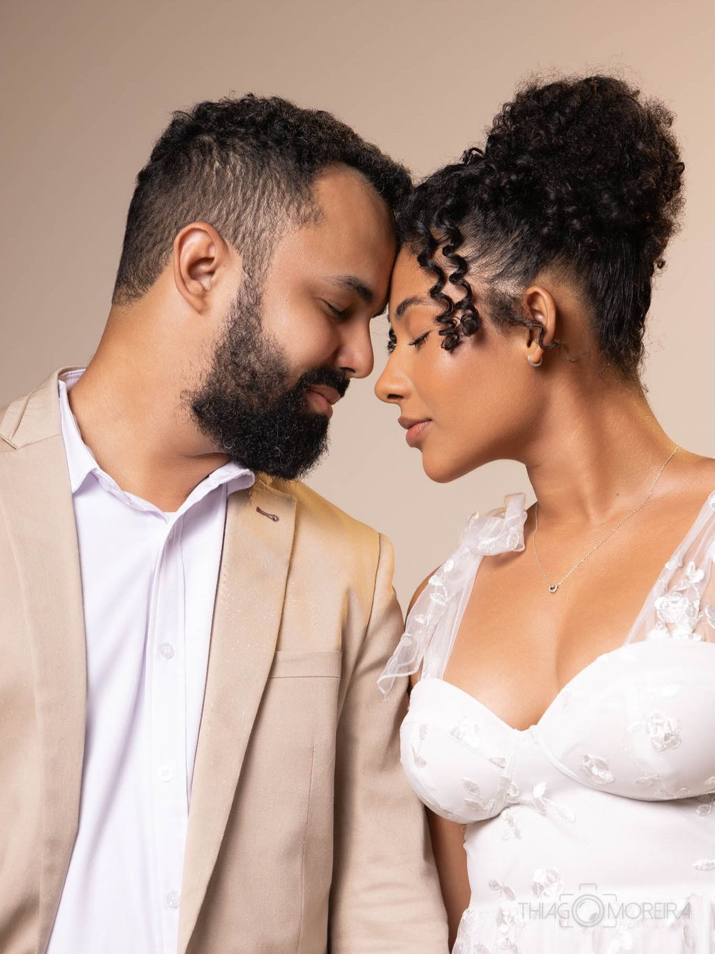 Casal de testa colada em ensaio de estúdio, ele de terno claro e ela de vestido branco rendado, sobre fundo bege